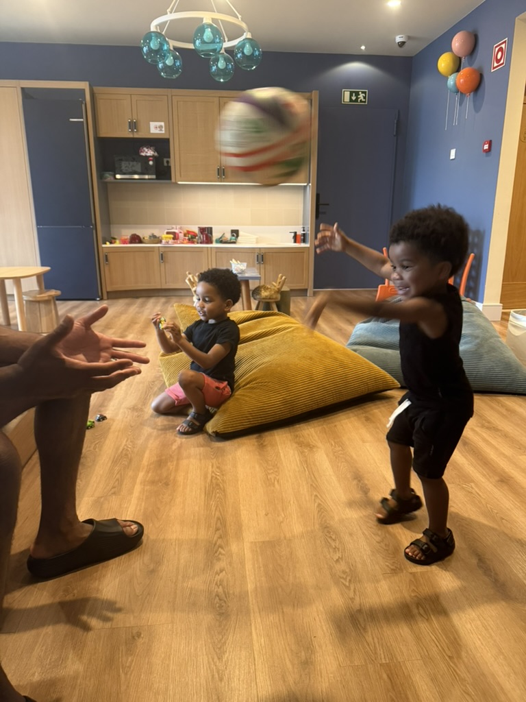 Two boys throwing a ball in a playroom with bean bags and an adult's hands reaching for it