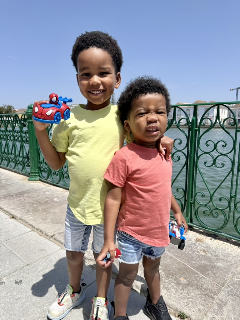Two young boys holding toy cars beside a green railing along the river in Tavira