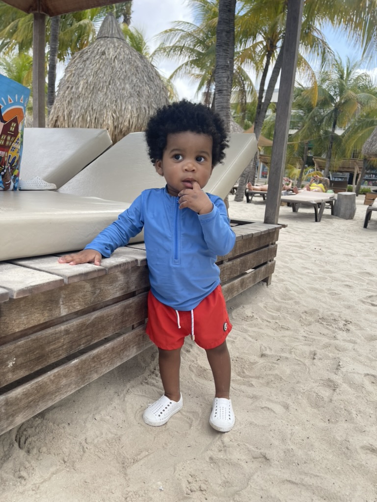 Toddler in a blue rash guard and red shorts standing beside a beach daybed under palms