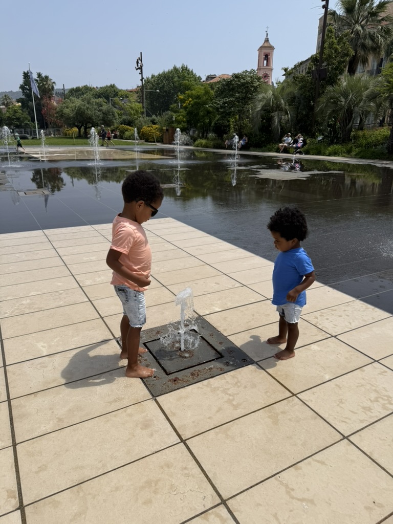 Two young boys standing barefoot over a small ground jet at a Nice splash pad with a bell tower behind them
