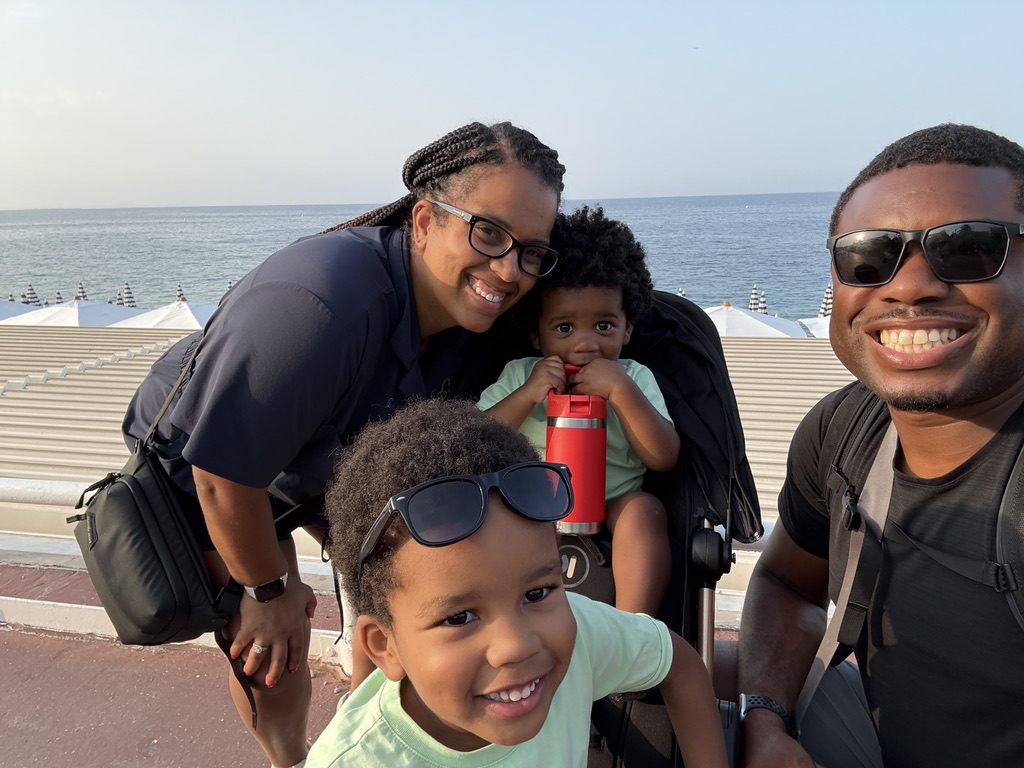 A family selfie of two parents and two young boys on the Promenade des Anglais with the sea behind them.