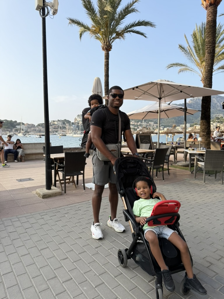 Father with two young children — one in a backpack carrier and one in a stroller — on a palm-lined waterfront promenade with boats and mountains behind