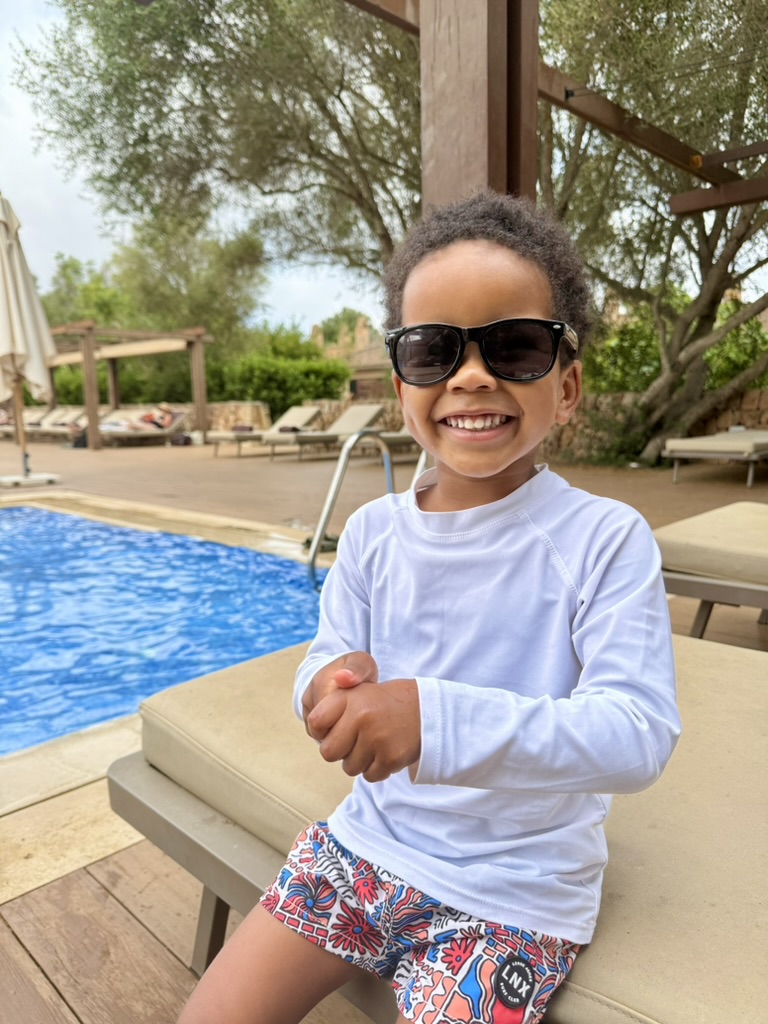 A smiling child in sunglasses and a white rash guard sitting on a lounger beside the pool