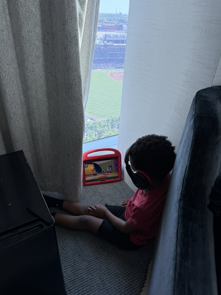 Preston sitting on the hotel room floor with headphones and a tablet beside the window over the ballpark