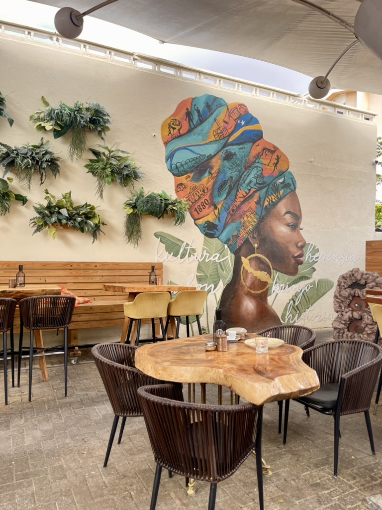 Shaded cafe patio with a mural of a woman in a colorful headwrap and hanging plants