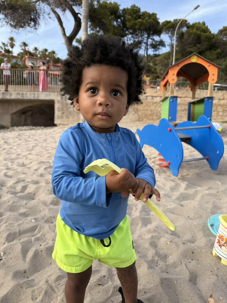 Toddler in a blue rash guard holding a sand tool near the beach playground back at Sant Elm