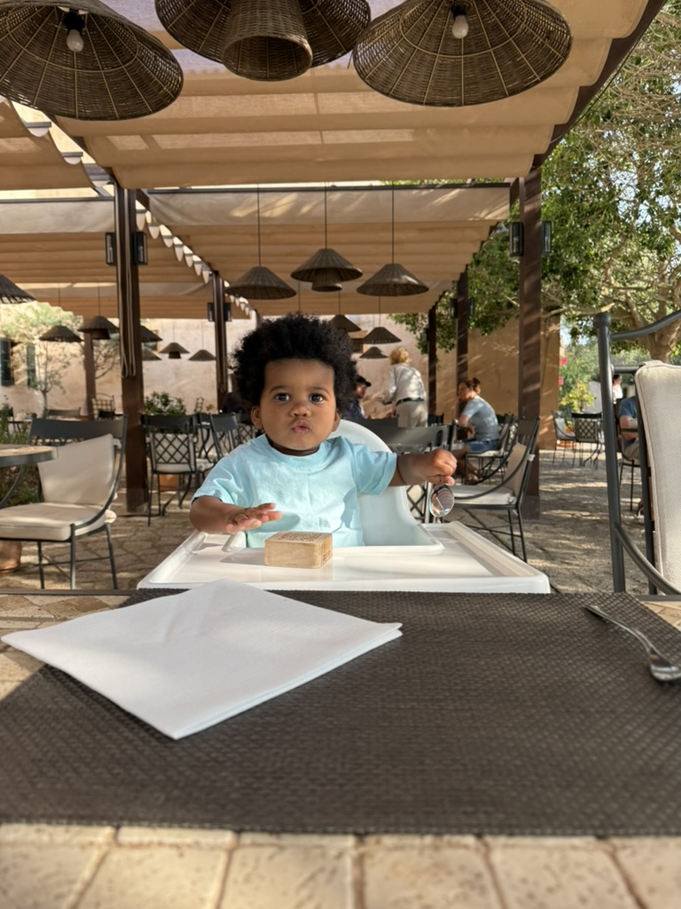 A toddler in a high chair with a piece of bread at a shaded terrace table under woven pendant lamps at Zoëtry