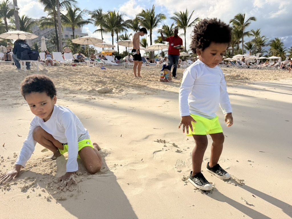 Two boys in white rash guards digging in the sand on Cable Beach with palms and umbrellas behind them