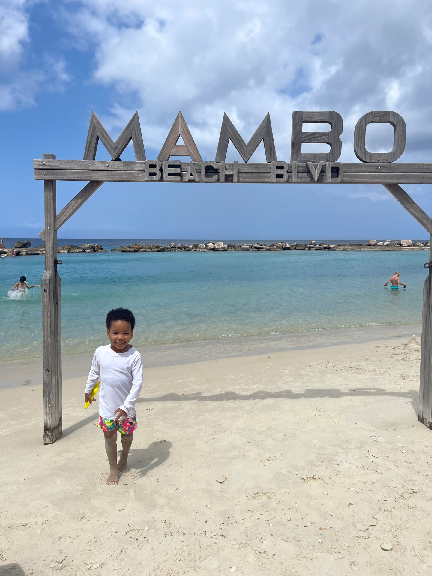 A toddler standing under the Mambo Beach Blvd sign with calm turquoise water behind him