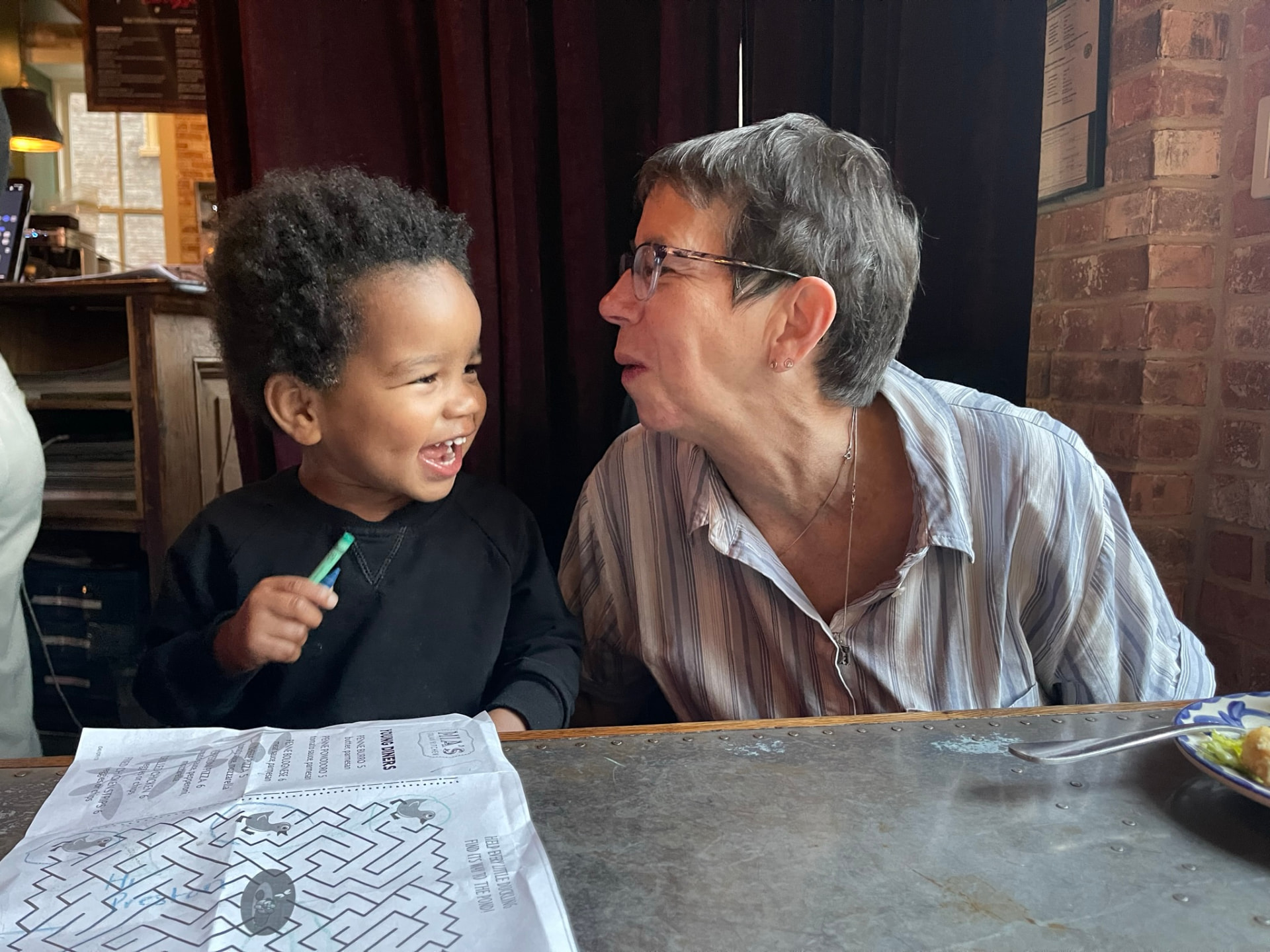 Toddler laughing with his grandmother at a restaurant table with a kids’ activity menu