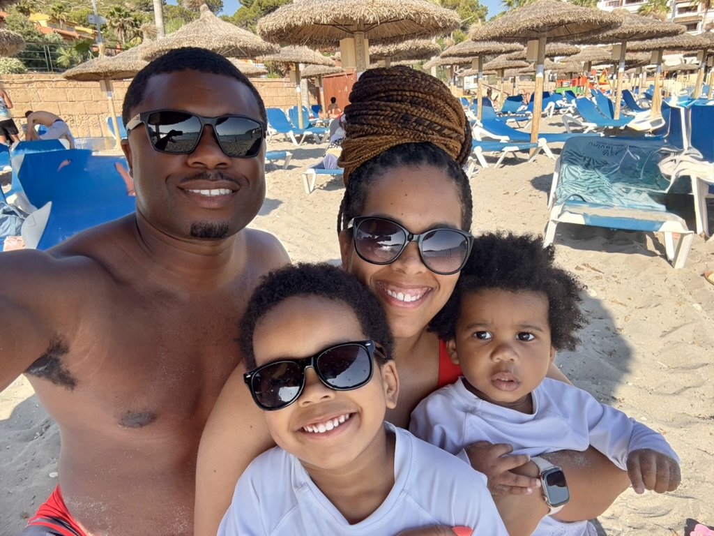 Family selfie on Sant Elm beach — parents and two young boys in sunglasses and rash guards under the thatched umbrellas
