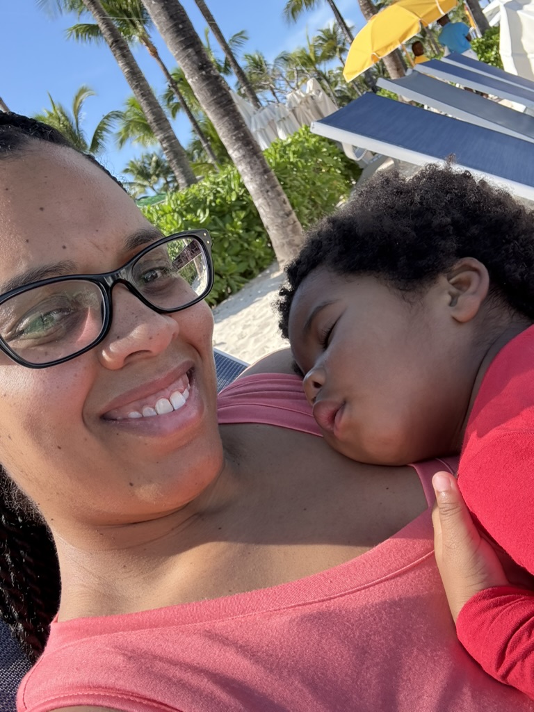 Mom smiling on a beach lounger with a sleeping toddler resting on her shoulder